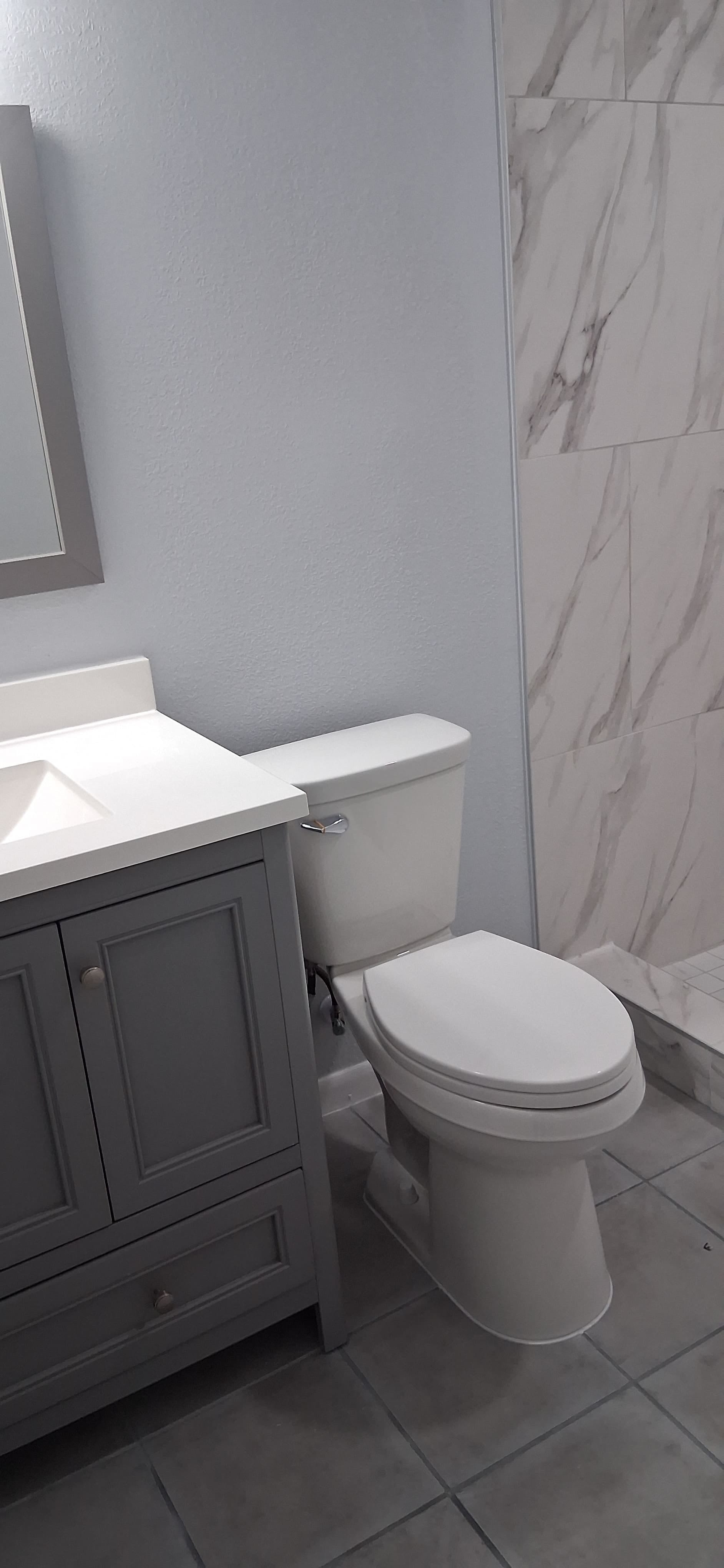 Bathroom with installed toilet, gray vanity, tile floor, and marble-look shower wall.