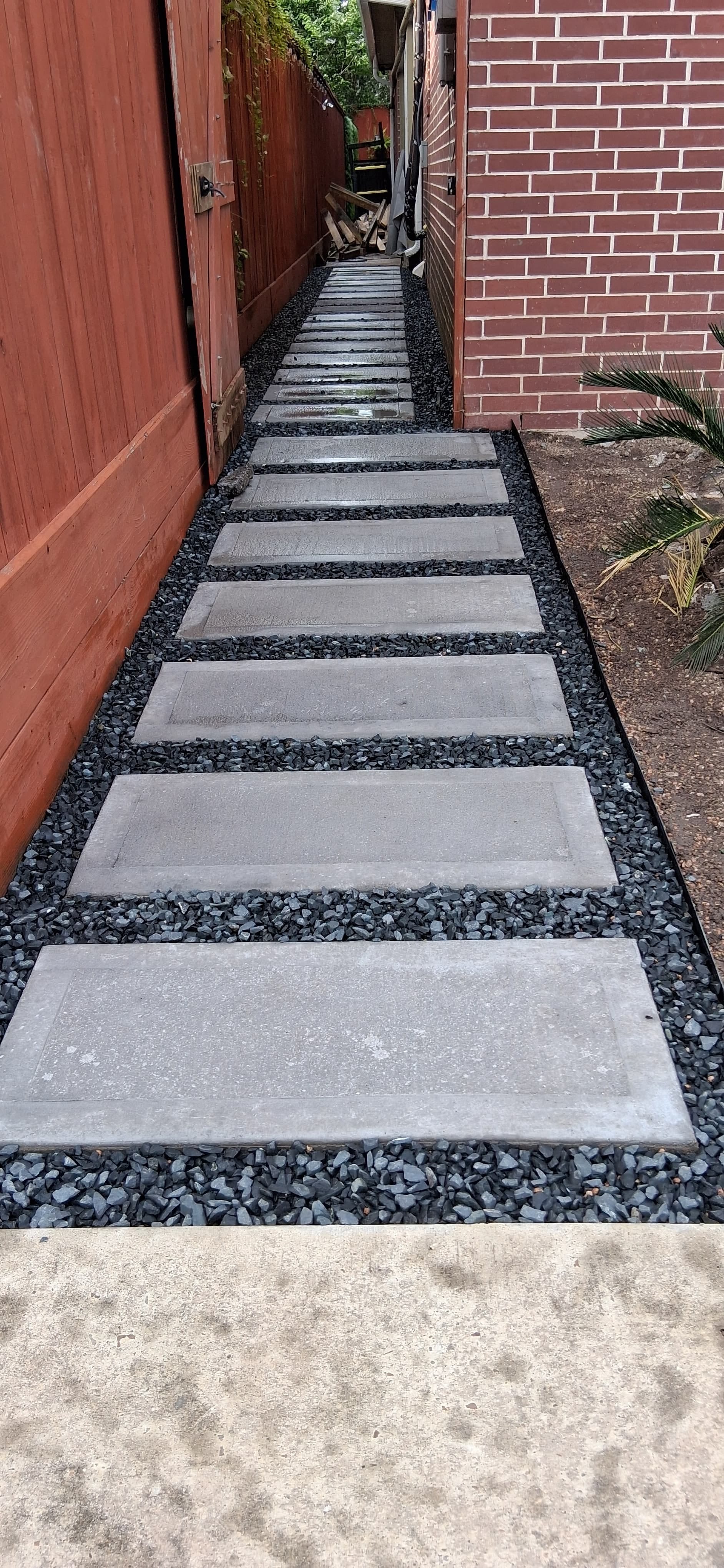 Finished concrete stepping-stone walkway with dark gravel between a fence and brick wall.