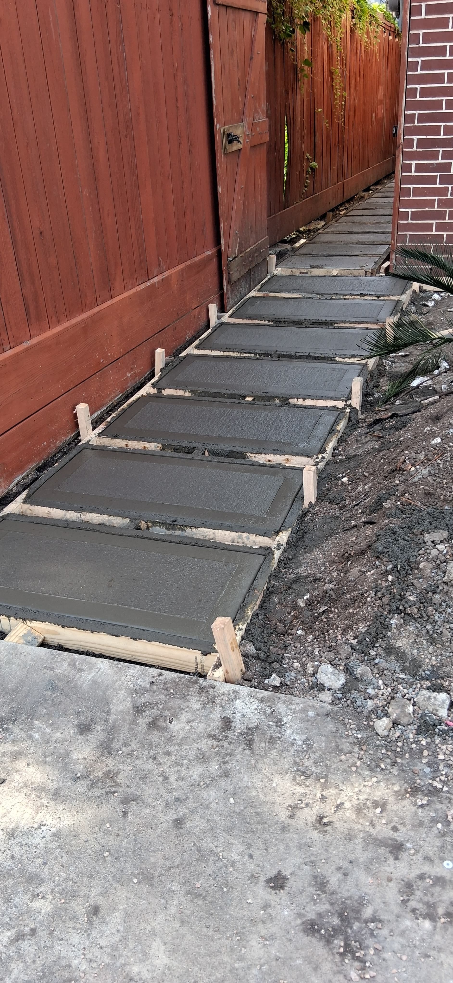 Walkway installation in progress with form boards, gravel, and concrete pads beside a fence.