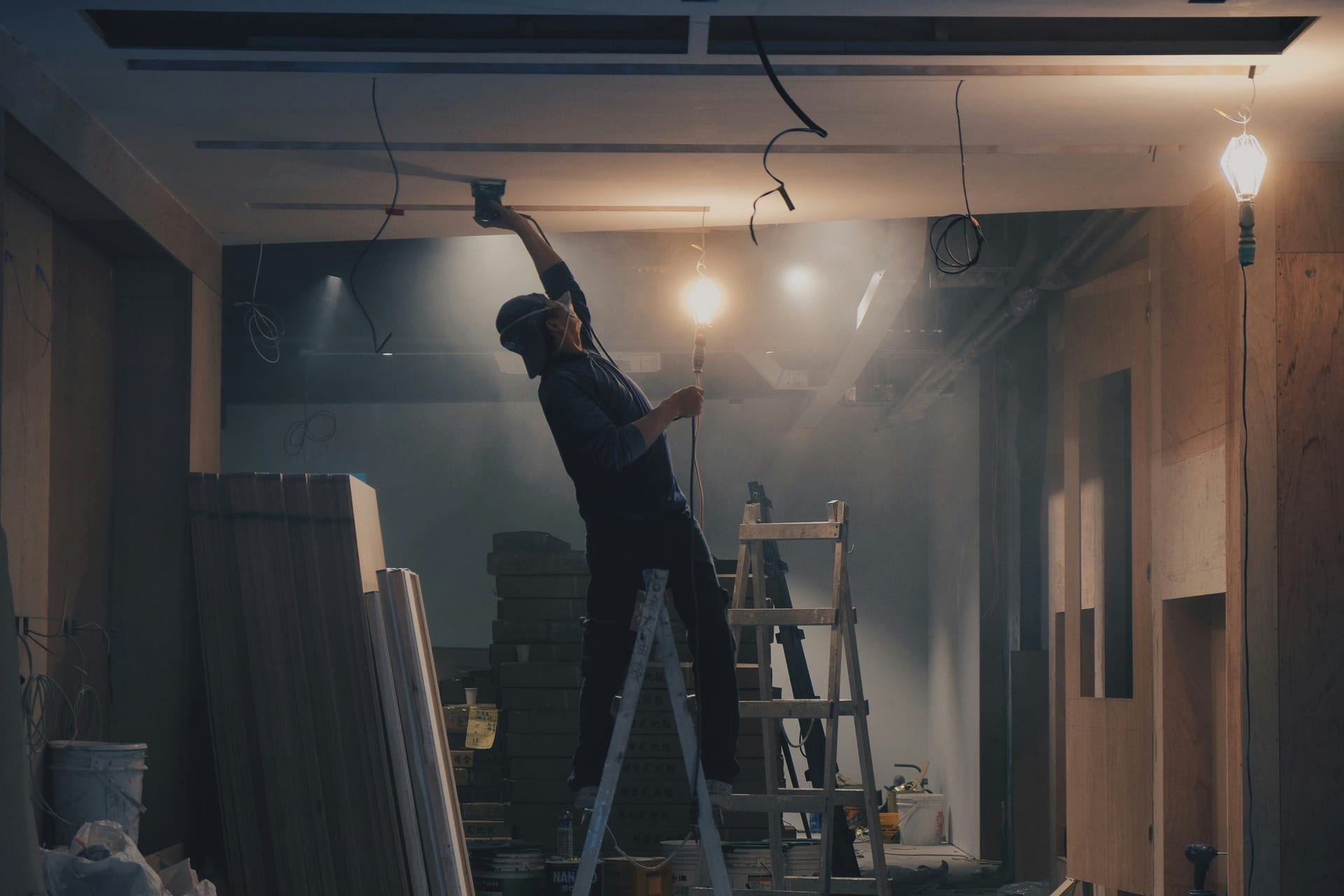 Worker on a ladder making an overhead repair with a drill and work light.